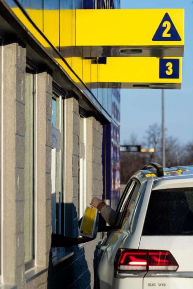 Yellow signs with numbers ‘2’ and ‘3’ above a drive-thru as a person in a white car grabs a yellow bag.